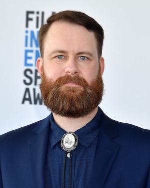 A bearded white man in a blue suit and bolo tie stands in front of a white background.