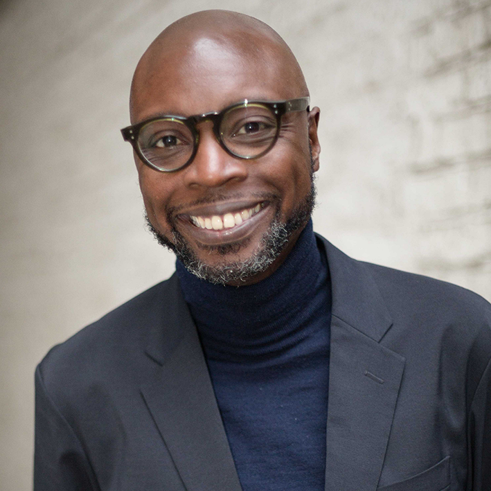 Derren Lawford, a black man wearing glasses and a suit, looks directly at the camera and smiles.