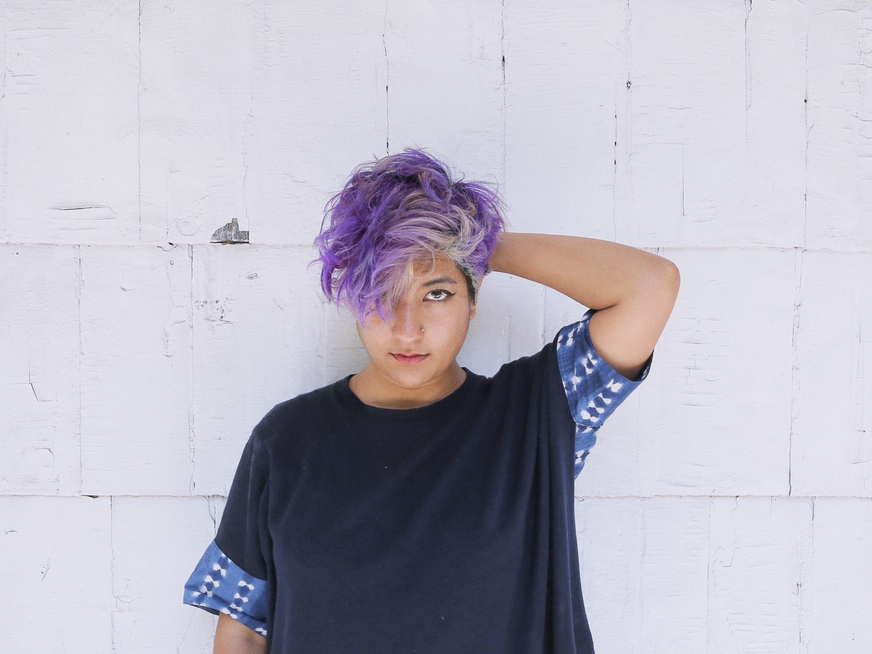 A South Asian woman with short, textured purple hair and a blue shirt with tie-dyed cuffs stand in front of a white wall, lifts one arm behind her head and looks directly to camera.