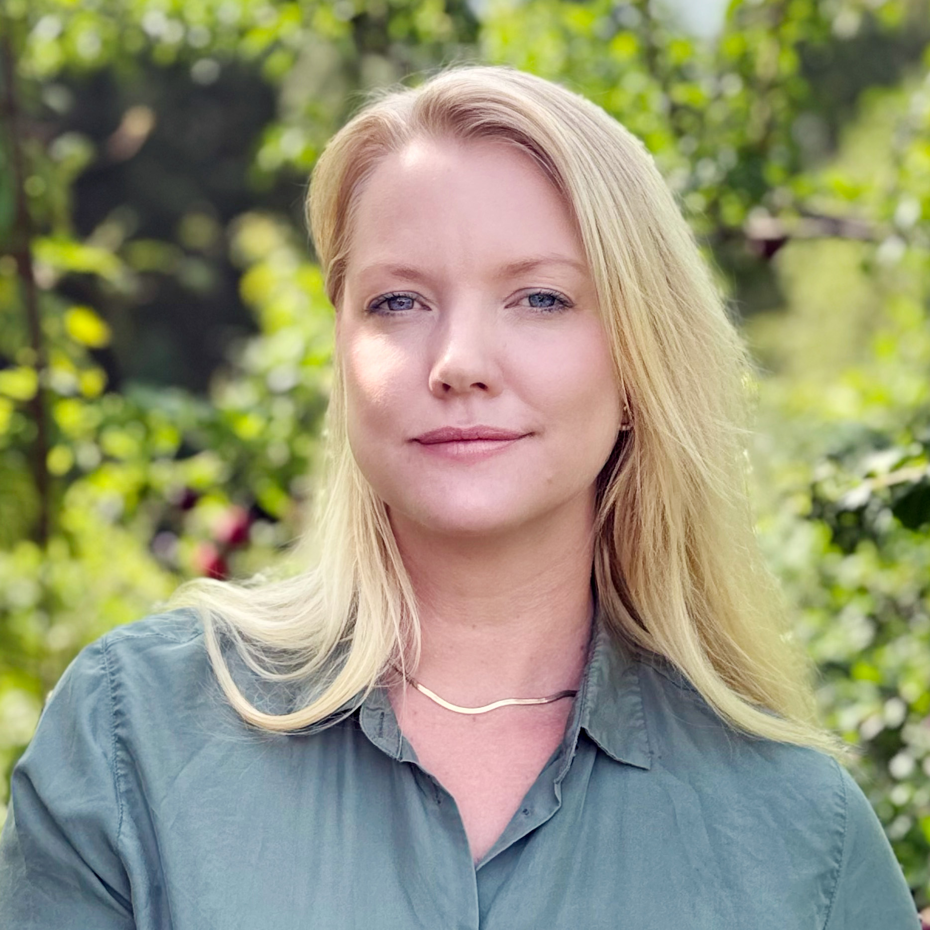 Blonde woman looks into the camera, blurry trees in the background.