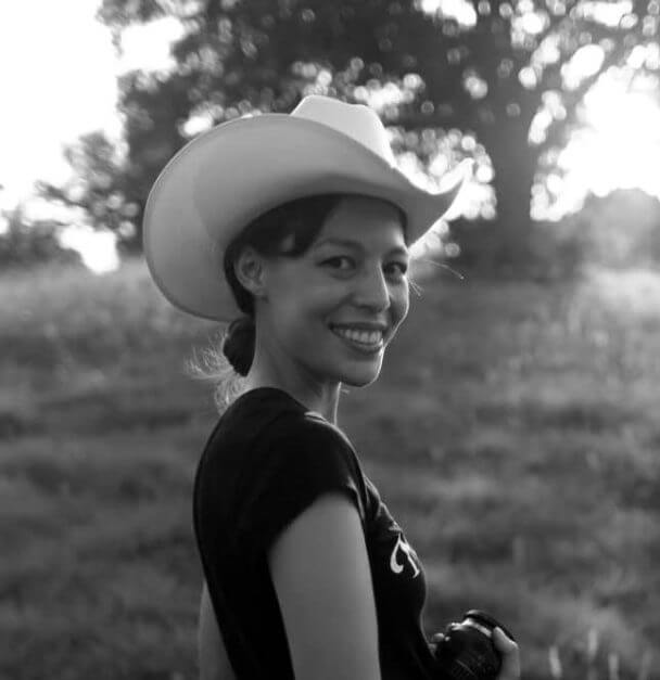 Sonia Kennebeck looking directly at the camera. She is standing in a green field with a tree in the background and is holding a photo lens. Black and white portrait