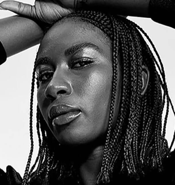 Sabaah Folayan looking directly at the camera. She is wearing blue braids and red lipstick. Black and white portrait.