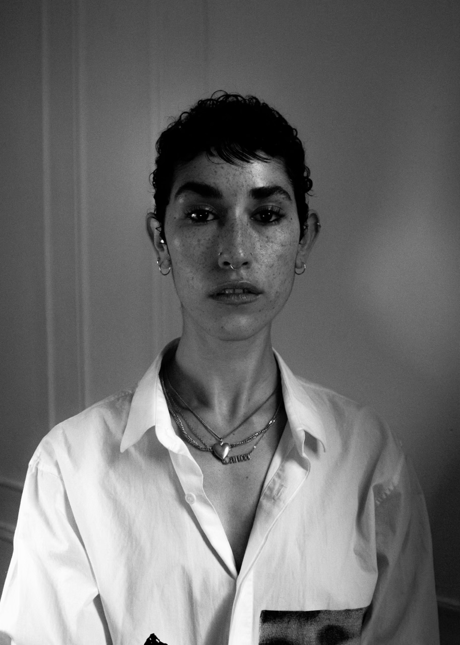 A black & white image of a mixed-race Black and white person with a dark pixie haircut, wearing a white button up shirt and layered necklaces. 