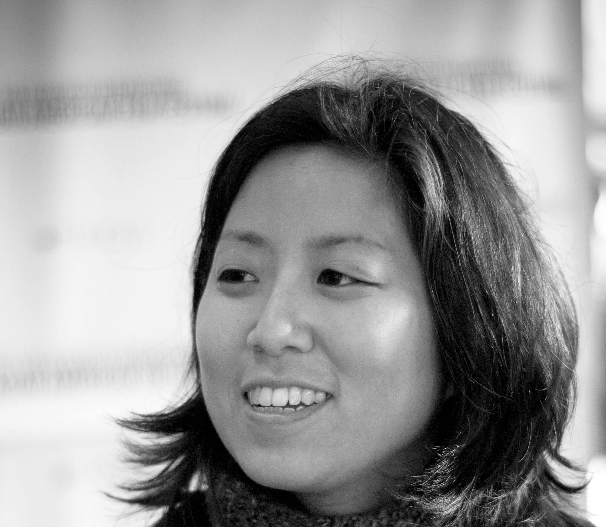 Grace Lee has short, dark hair, wears a coat and a scarf, looks away from the camera and smiles. Black and white portrait.