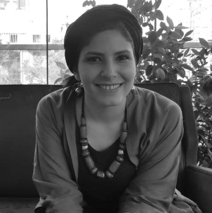 A black and white headshot of filmmaker Sara Khaki. She wears a scarf on her head and a large beaded necklace. She is seated, smiling, looking directly at the camera, leaning toward the photographer/viewer. 