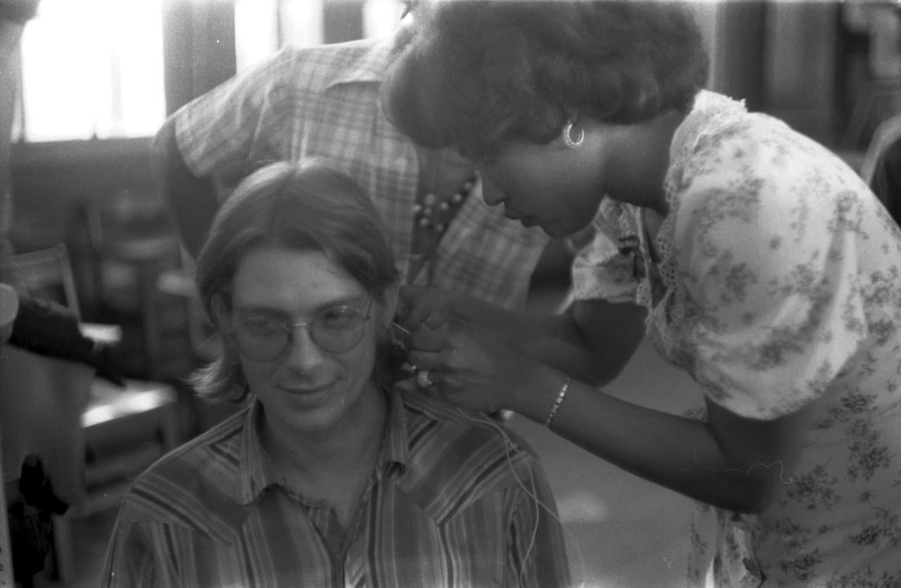 Still image from Dope Is Death. A white man gets his ear pierced by a black woman with a needle and thread.