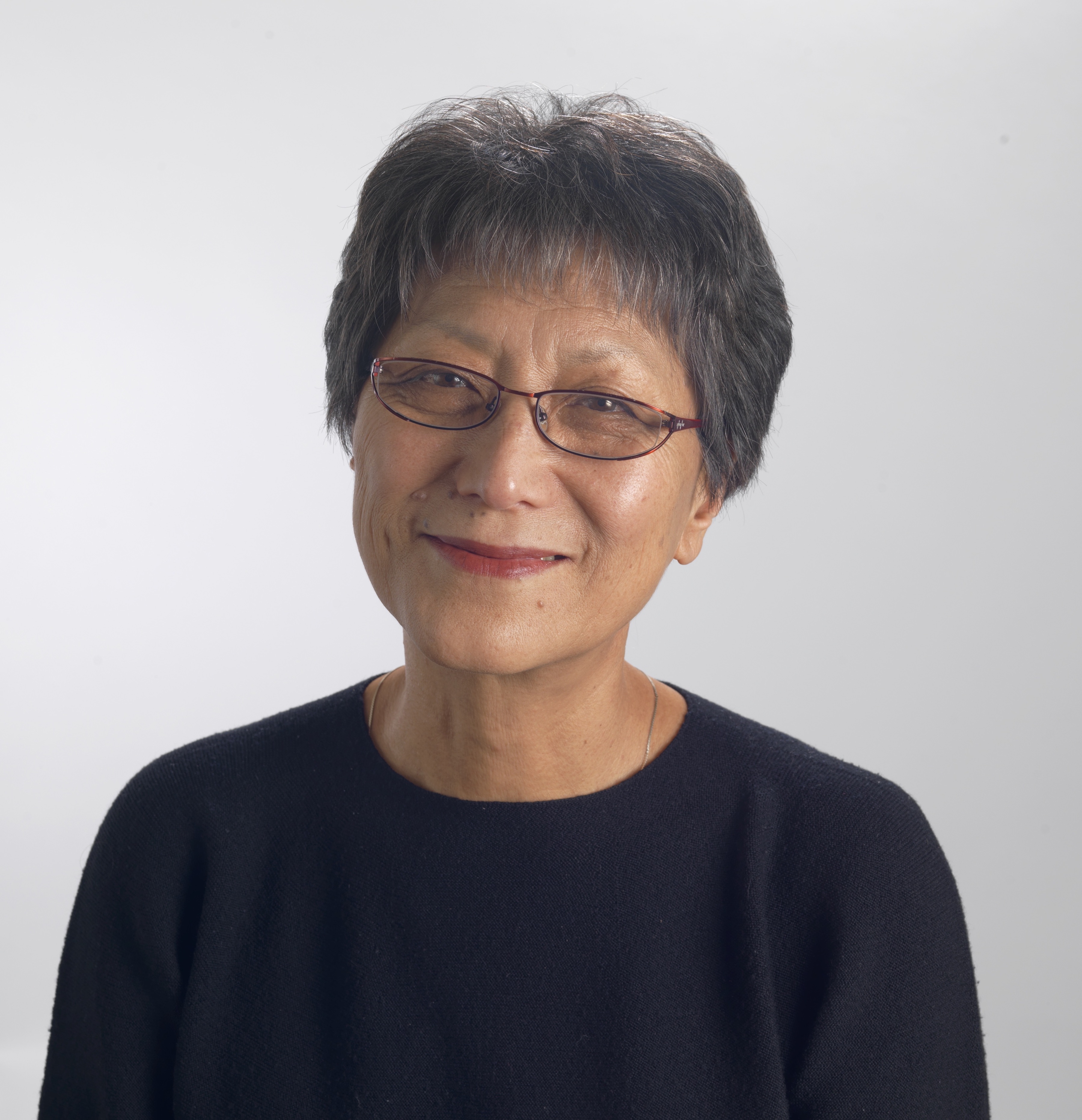 Headshot of Deann Borshay Liem, she wears a black round neck shirt, thin frames, and short hair. She smiles at the camera. 