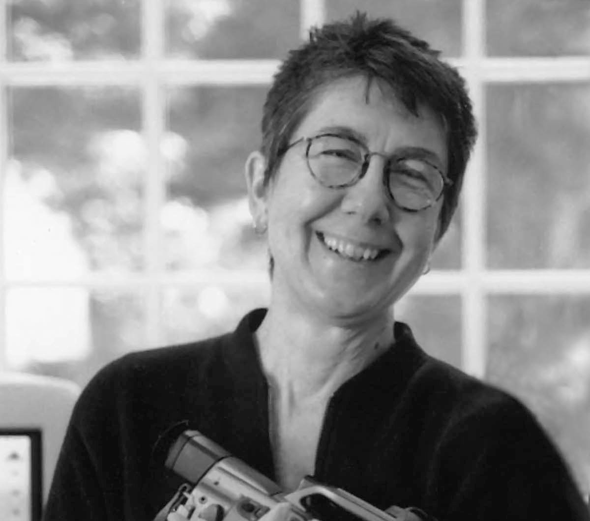 Julia Reichert wearing a dark blouse and holding a camera in her arms. She is smiling, has short hair and circled frames. Black and white portrait.