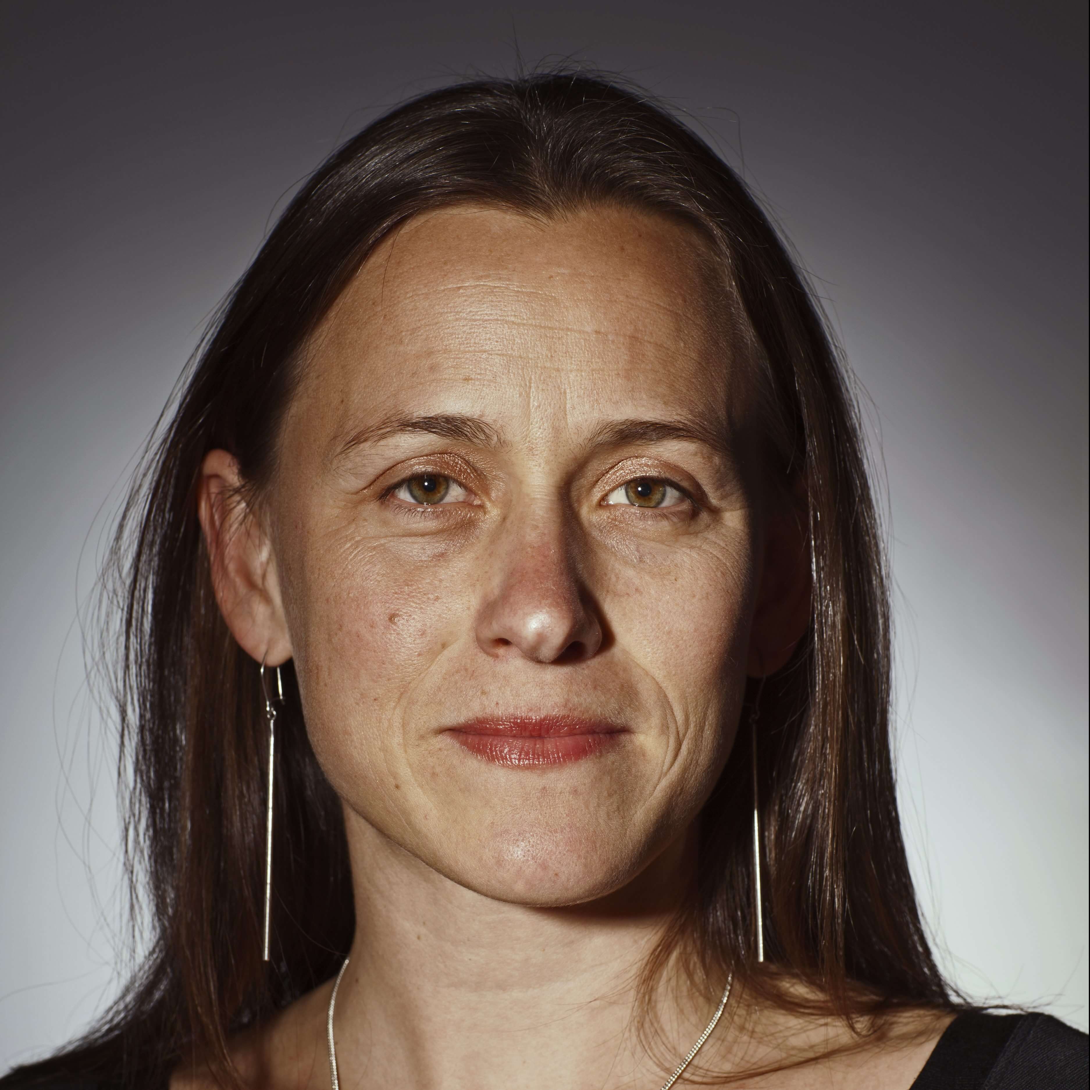 Natalia Almada smiling slighlty, wearing a black top, long earrings, and a ring on a necklace. Her hair is long and brown, and she wears shimmery eye shadow with an orange-red lipstick. She wears a black circled neck top, long earrings and a ring in a necklace.