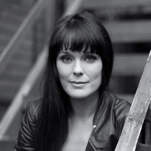 Michelle Latimer sitting on a fire escape stair. Her hair is long and pulled to her left, and she wears a jacket. Black and white portrait.