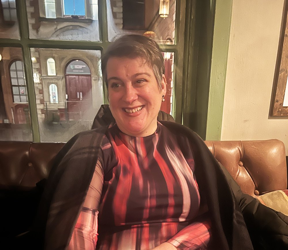 Suzanne Alizart headshot. Suzanne is a white woman with short brown and grey hair. She is wearing a dress with a deep red and plum pattern and is smiling warmly. 