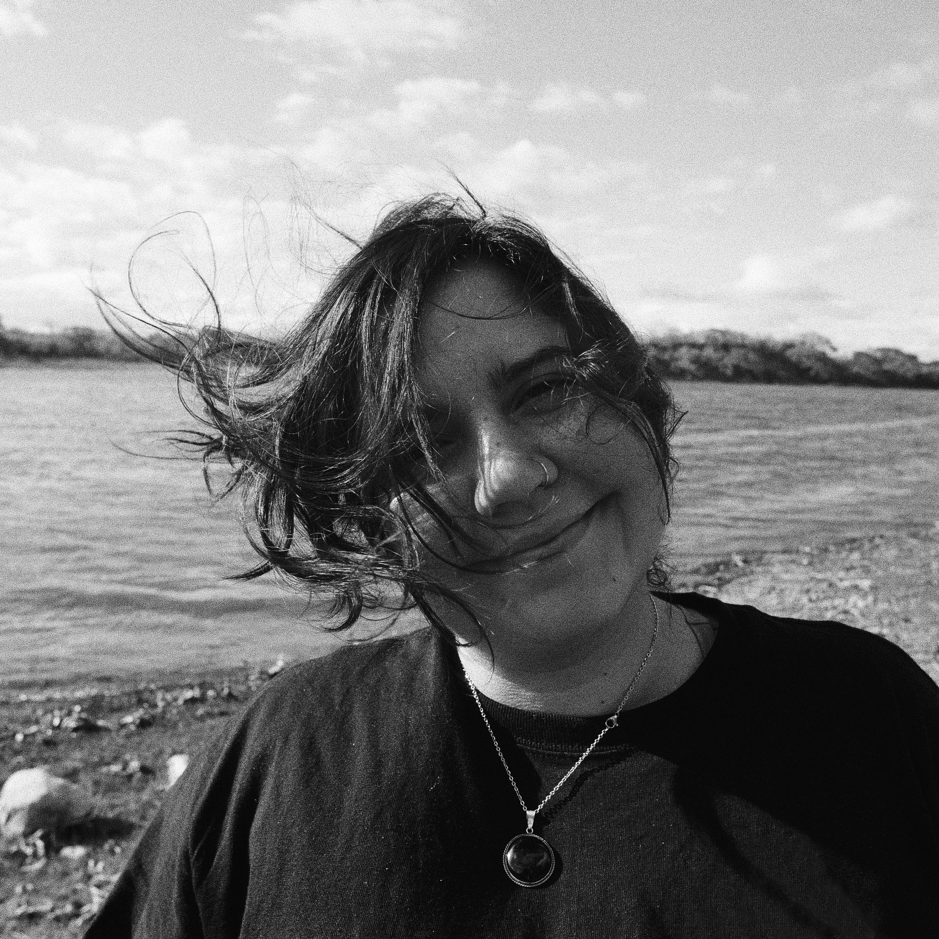 A woman, Amada Torruella, smiles and looks to the camera as wind blows her hair. In the background we can see a cloudy sky and a body of water.