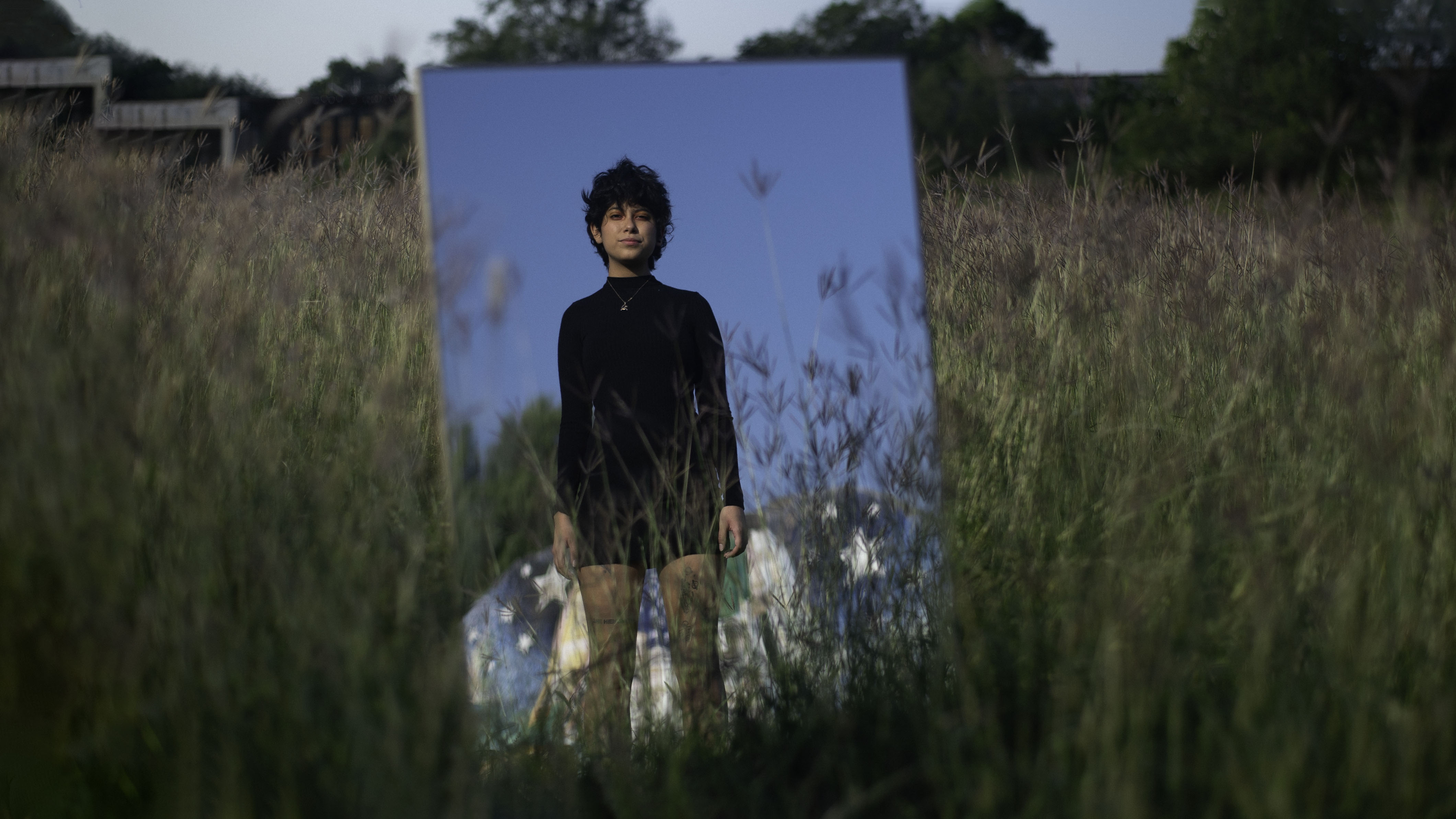 Photo of Silvia Castaños’s reflection on a mirror. She is wearing a long sleeve black dress in a field.