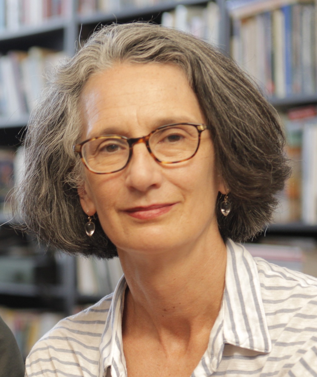 Janet Goldwater looking directly at camera. She is wearing eyeglasses and earrings. The background of bookcases is out of focus. Black and white portrait. 