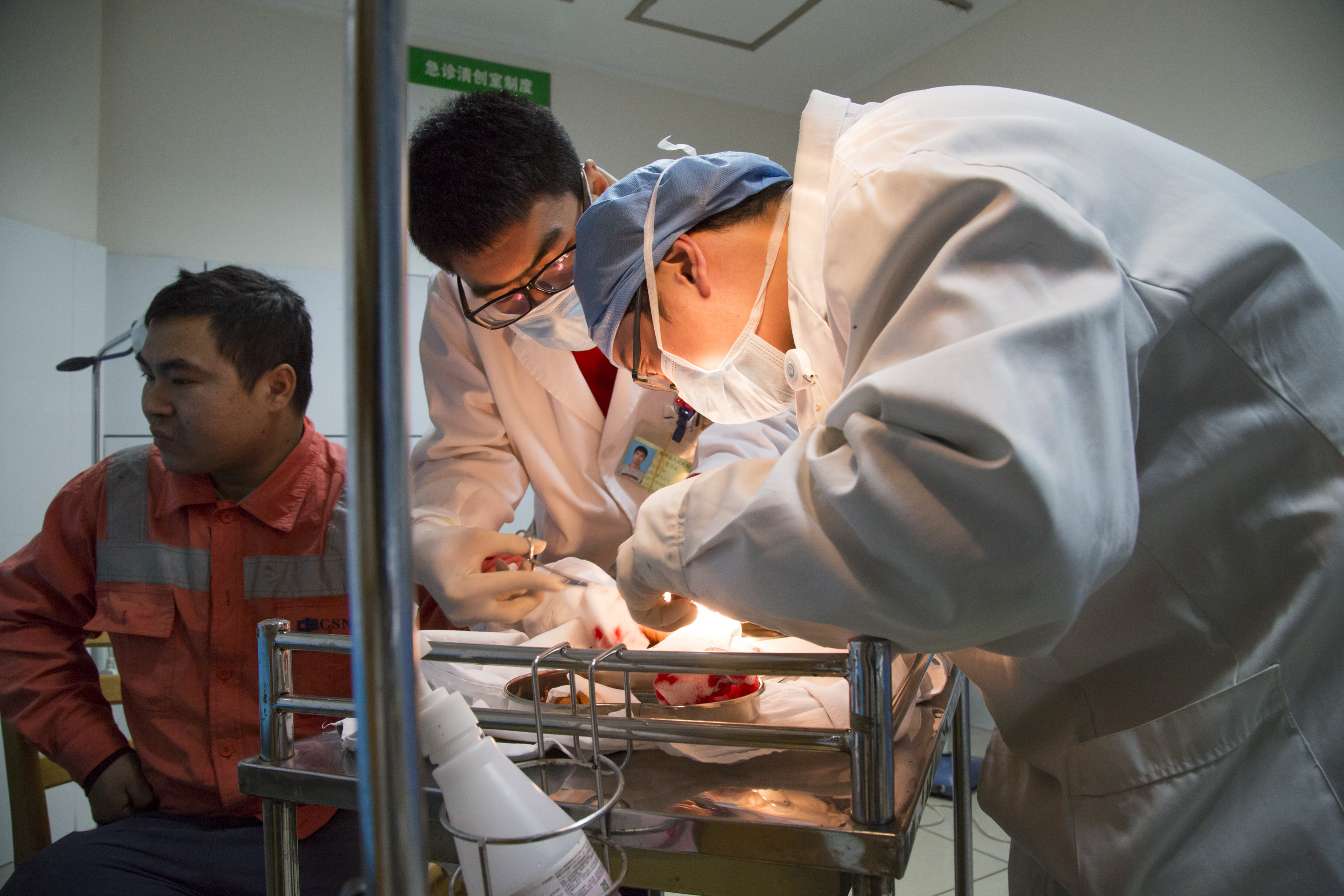 Still from Dear Mother, I Meant to Write About Death. Two medical staff in lab coats hover over a brightly lit tray of bloody gauze and a man in an orange jacket sits nearby.