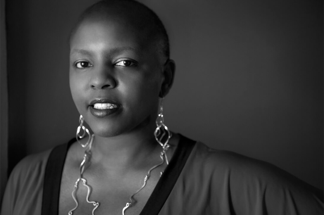 Yoruba Richen looking directly at the camera, with a short haircut. She wears matching earrings and a necklace, and a blouse. Black and white portrait.