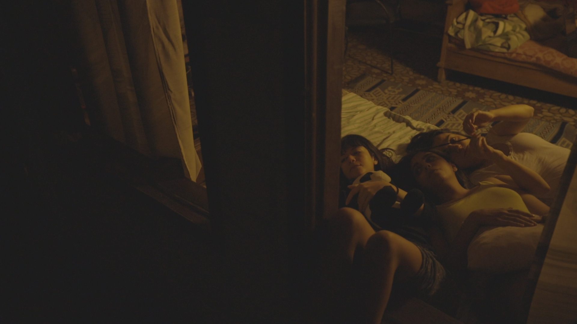 A still from House No. 7. Three young girls lay on a mattress in a dimly lit apartment.