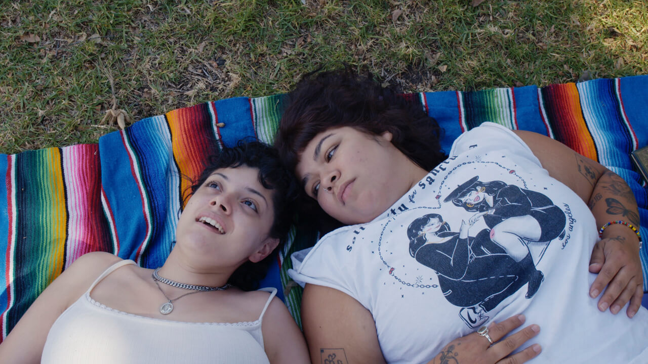 Two young women rest on a blanket on the grass while looking up.