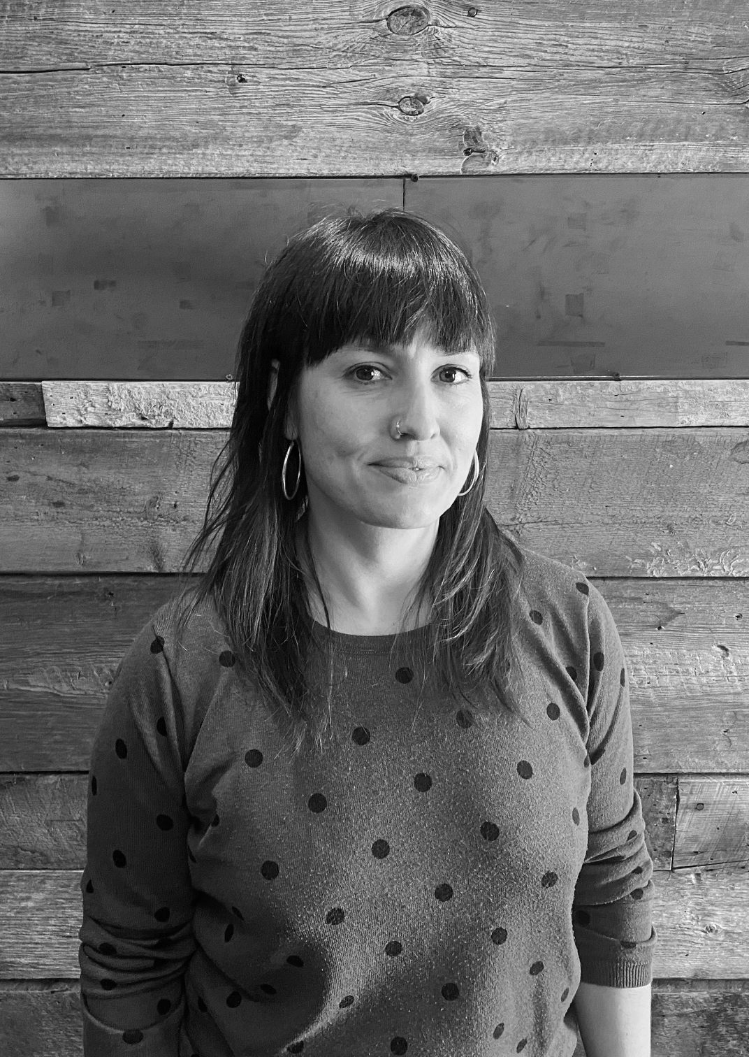 Rocío Barba Fuentes looking directly at camera. She is standing in front of a wooden paneled wall. She has hazelnut eyes, dark, medium length brown hair, a nose ring, silver earrings and is wearing a blue sweater with black dots. Black and white portrait. 