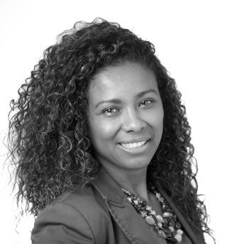 A headshot of a woman looking at the camera with curly medium length hair, wearing a blazer and a necklace