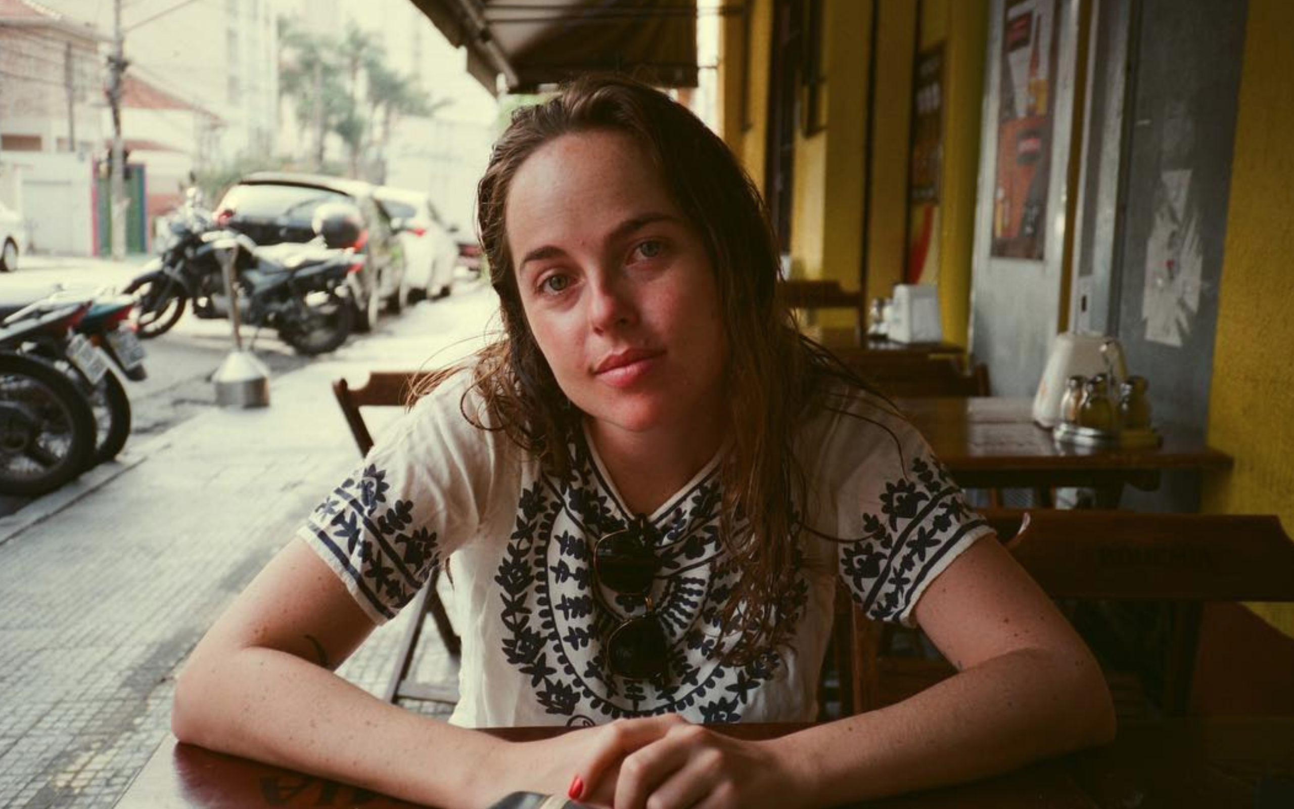 Portrait of Sofia Geld sat and folding her hands in a table, wearing an embroidered shirt, and smiling slightly at the camera, she has medium long hair. 