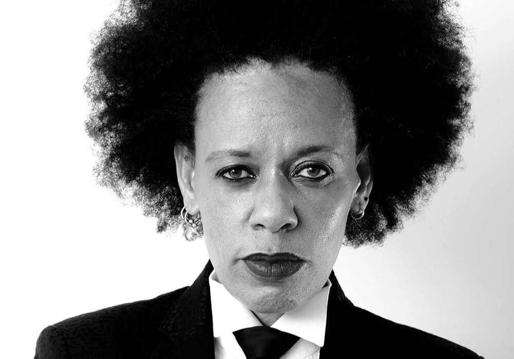 Medium image of Dr Bev in a Afro hairstyle, wearing a black tux and black tie, looking directly at camera.