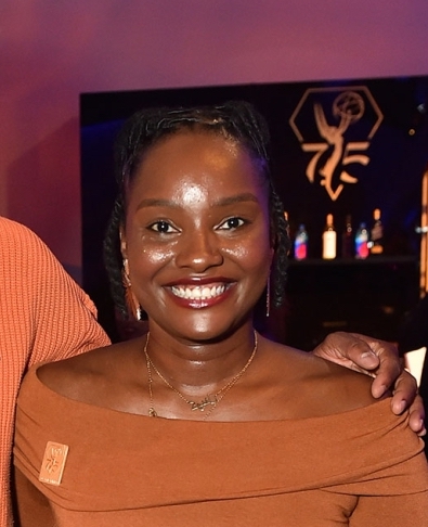 Brown skinned woman smiles at camera dressed in brown.