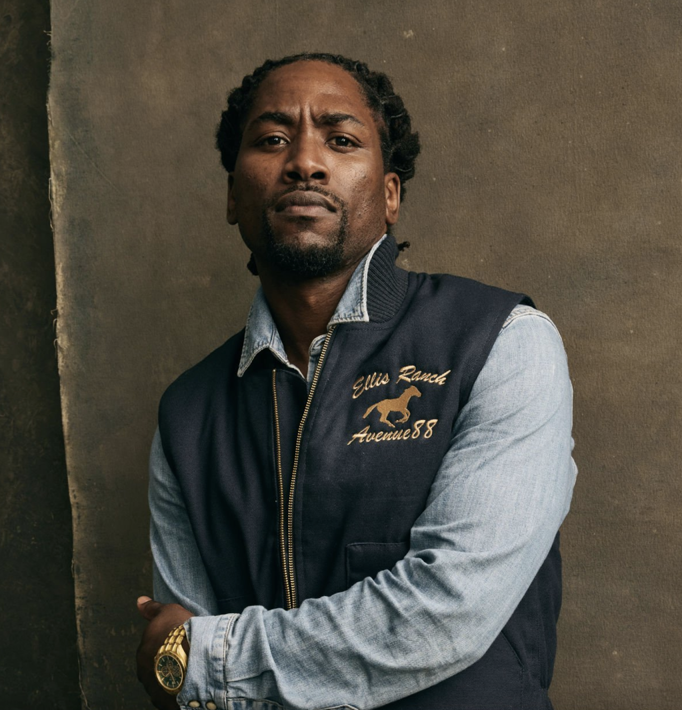 Headshot of JJ’88 wearing a denim shirt and navy blue vest against a brown backdrop.