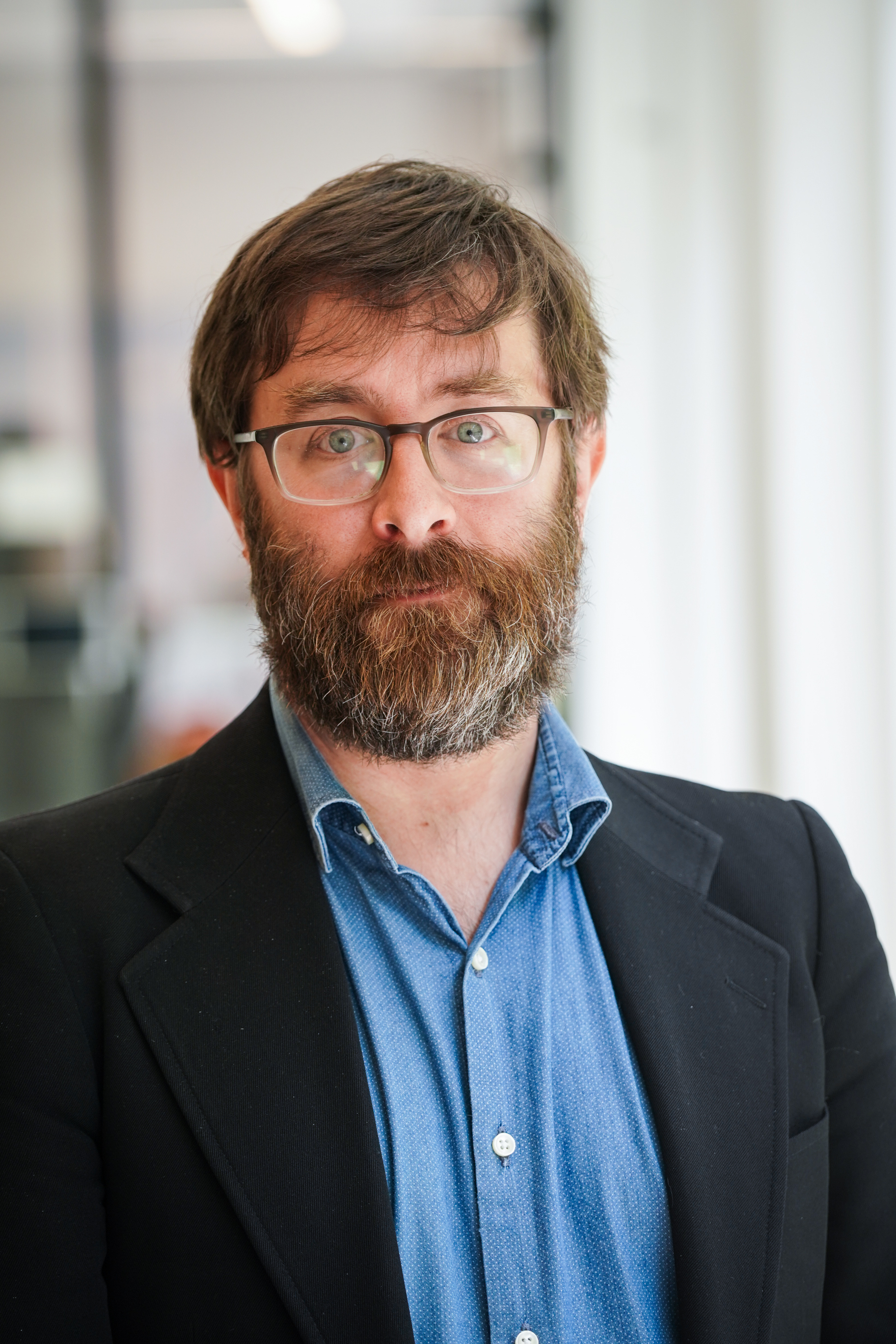 A caucasian man with brown hair, a salt and pepper beard, and blue eyes behind rectangular framed glasses looks direct to camera.