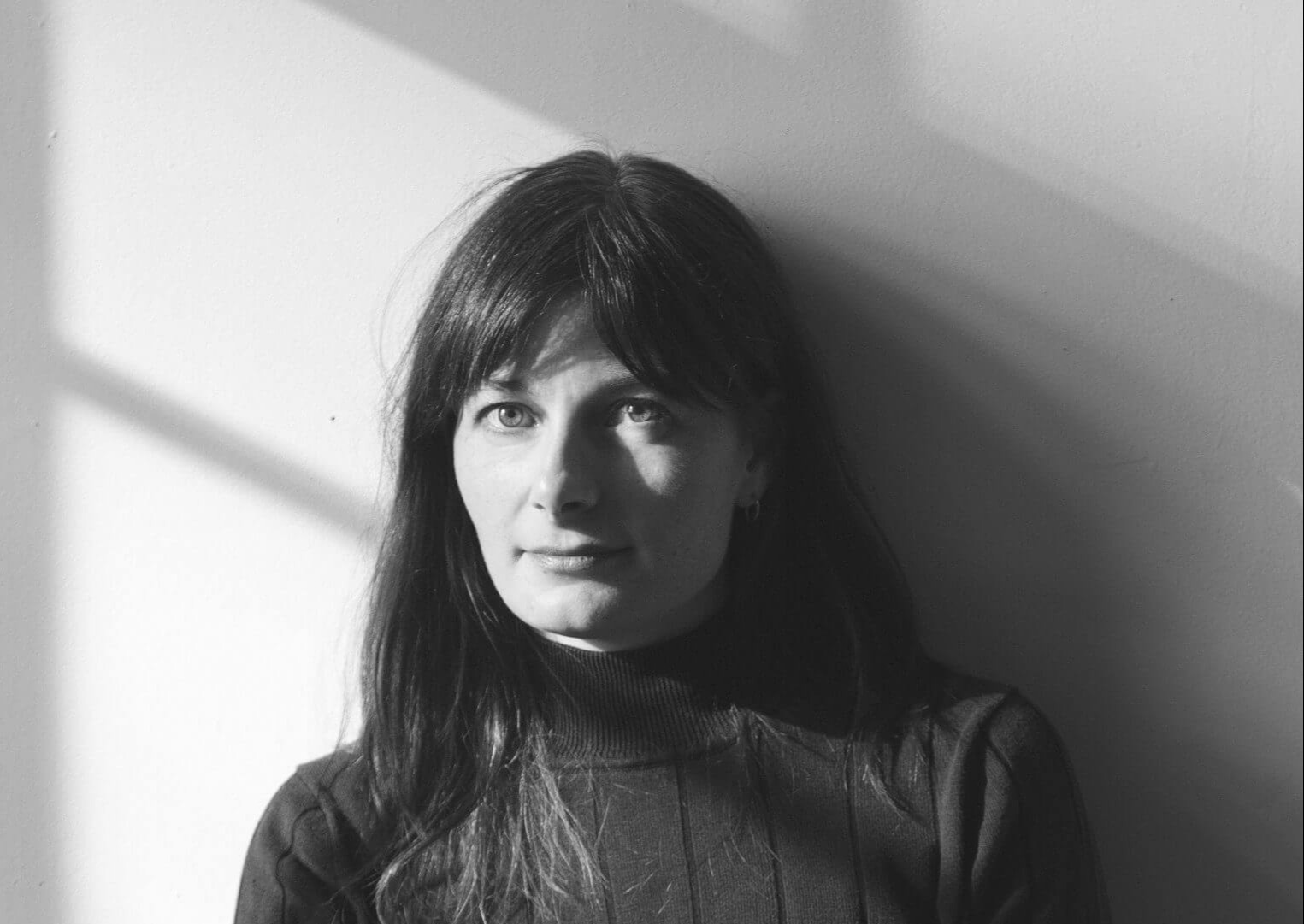 This is a headshot of Brett Story, a white woman with freckles and long brown hair and bangs. She is sitting at a table against a white wall, wearing a black turtleneck and jeans.