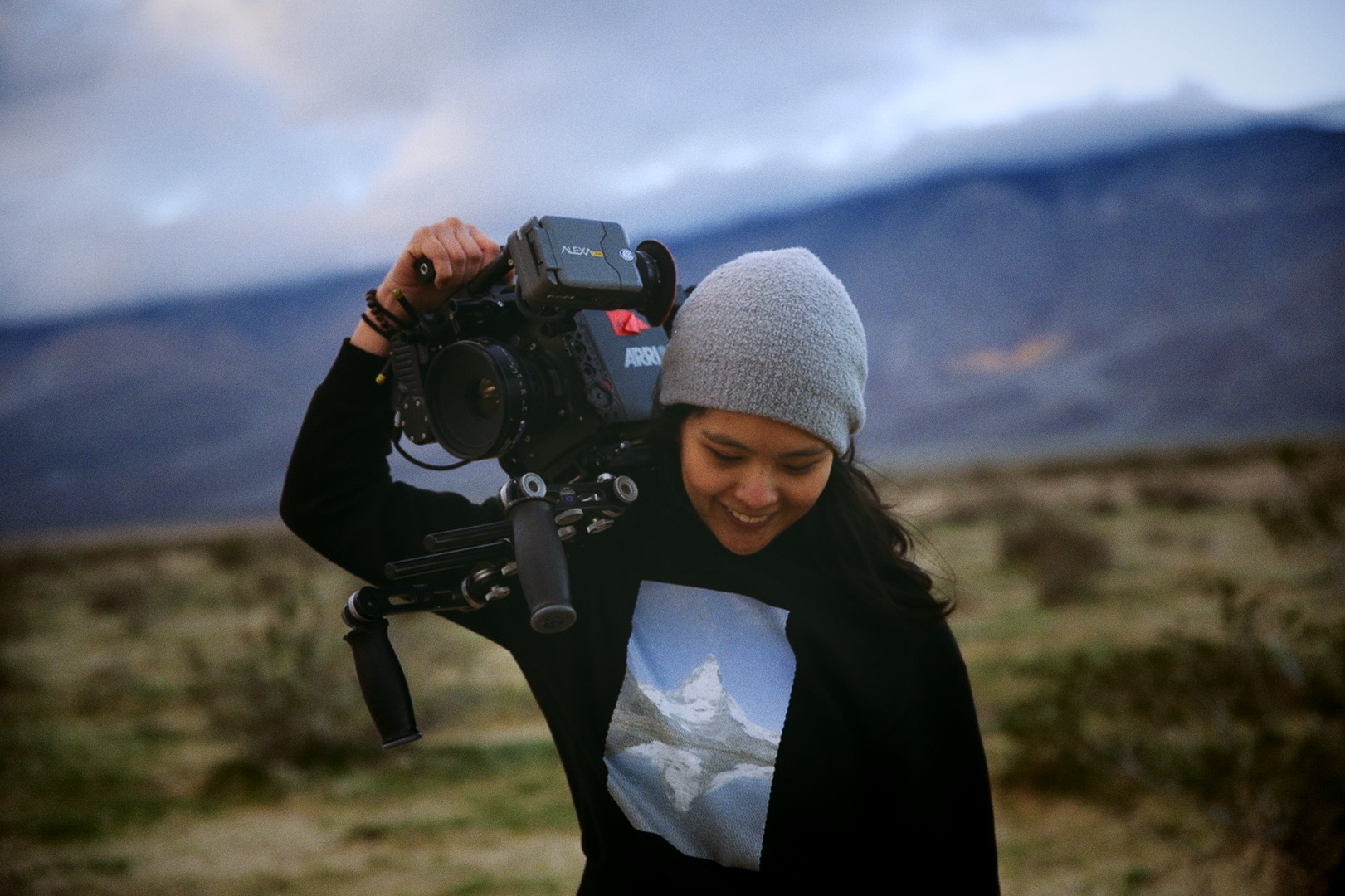 Portrait of a woman looking at the ground while holding a camera on her right shoulder, she wears a hat and a long sleeve shirt