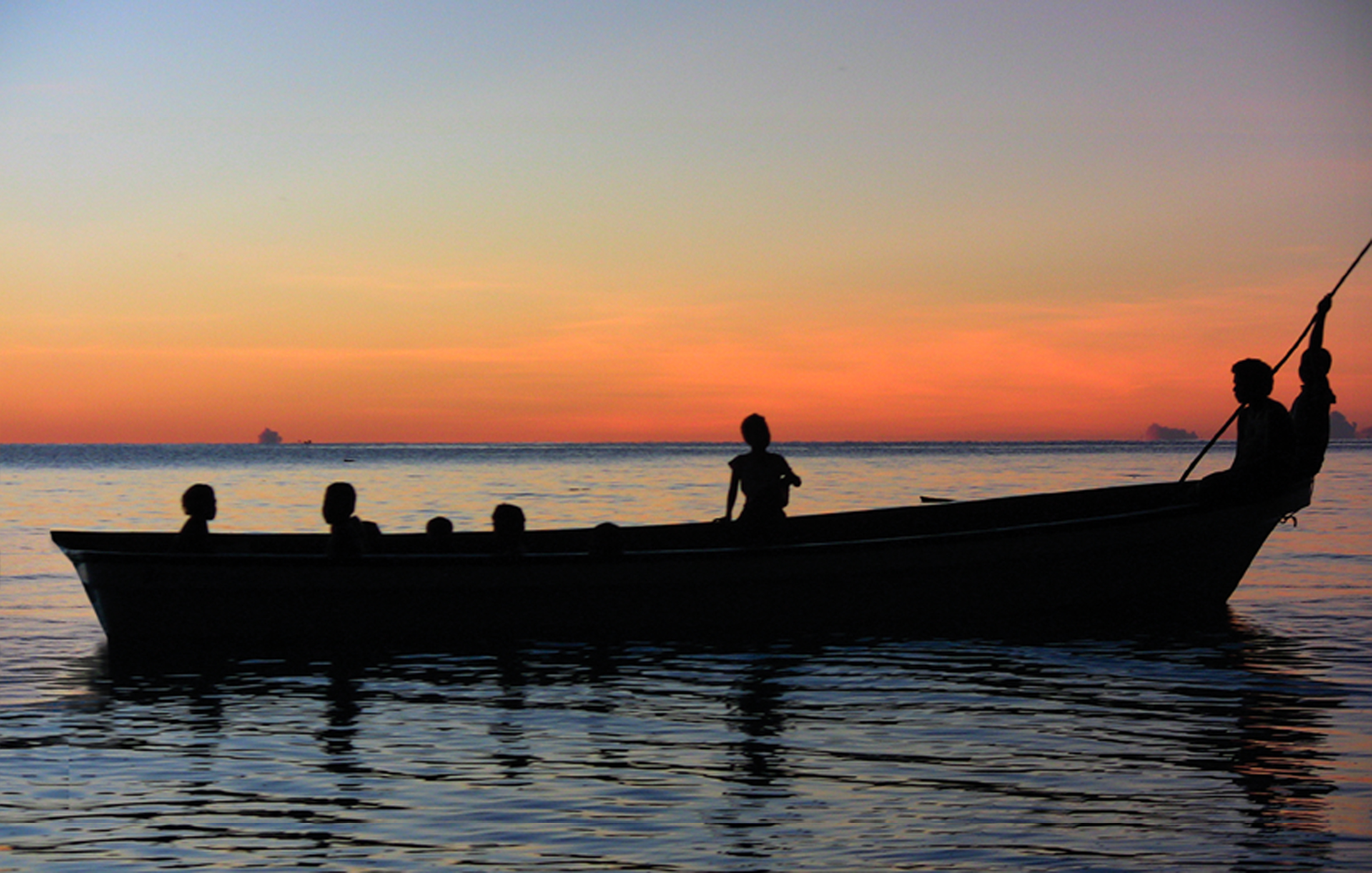 Still from Sun Come Up. A photo of four children sitting in a boat on the water. It is sunset, the water is dark blue and the sky features a vibrant sunset of yellows, reds, and oranges. Only the silhouette of the children on the boat is visible.