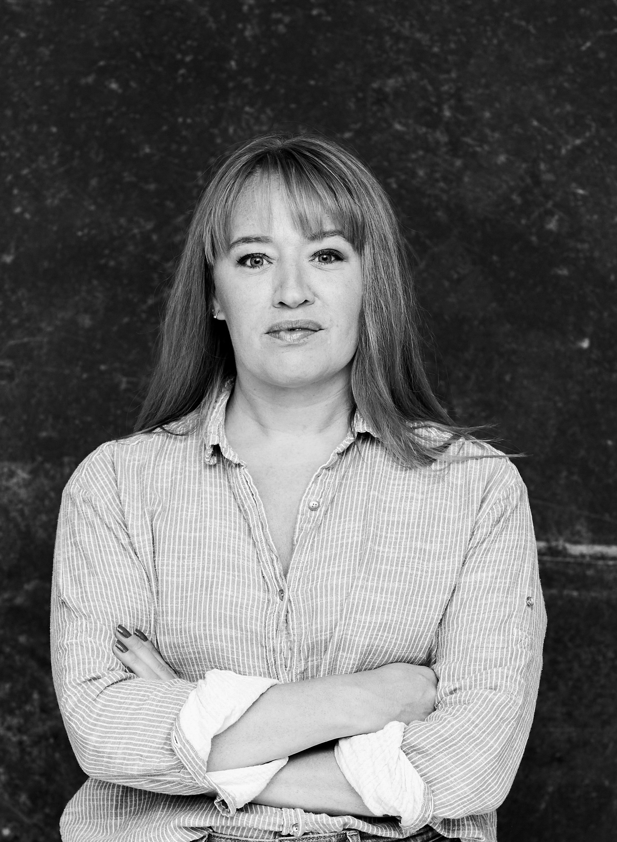 A black and white photo of a blondish woman with shoulder length hair and light eyes wearing a striped, collared shirt. Her arms are folded.