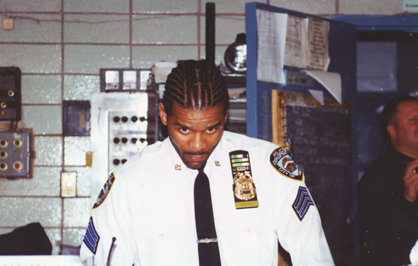 Corey Pegues, one of the highest ranking black executives in the NYPD, wearing his police uniform with many insignia and badges to indicate his seniority. his hair is braided and he looks up slightly toward the photographer while standing at a desk inside his precinct. 