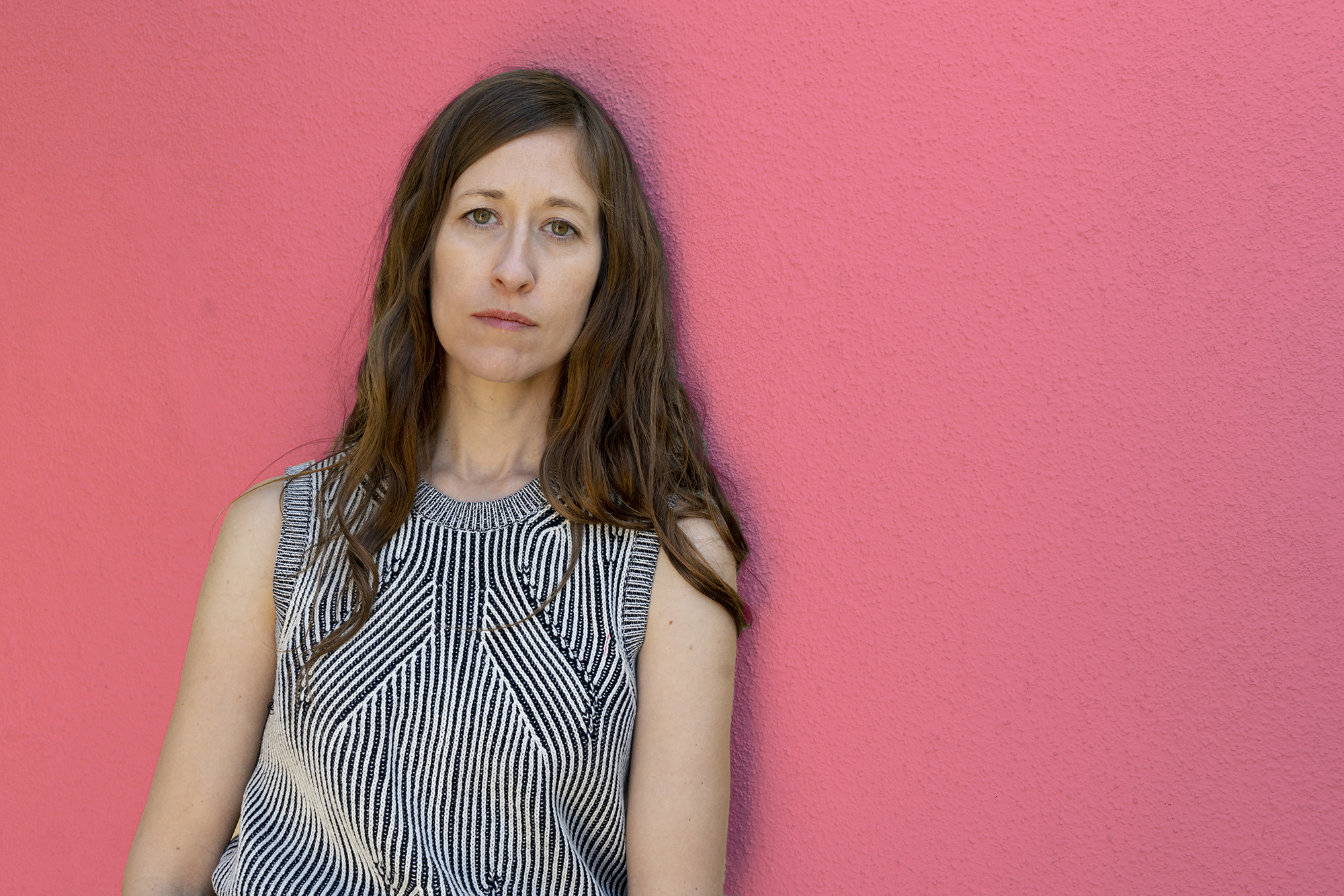 Alison O'Daniel looking directly at the camera. She wears a sweater tank top and leans against a pink wall. 