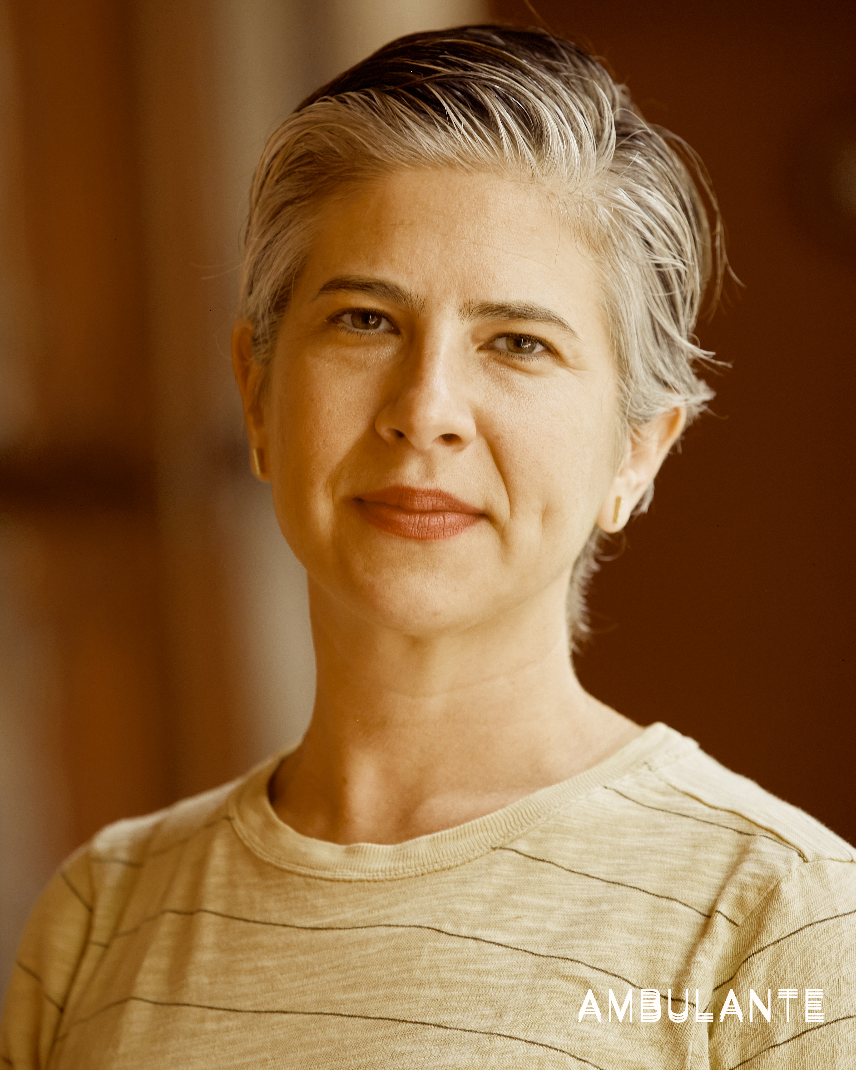 Emily Cohen Ibañez looking directly at the camera. She has short grey and dark brown hair and is wearing gold earrings, lipstick, and a striped T-shirt. 