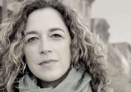 Kristi Jacobson looking directly ahead. She has shoulder-length hair and wears a leather jacket and scarf. Buildings are seen in the background out of focus. Black and white portrait.