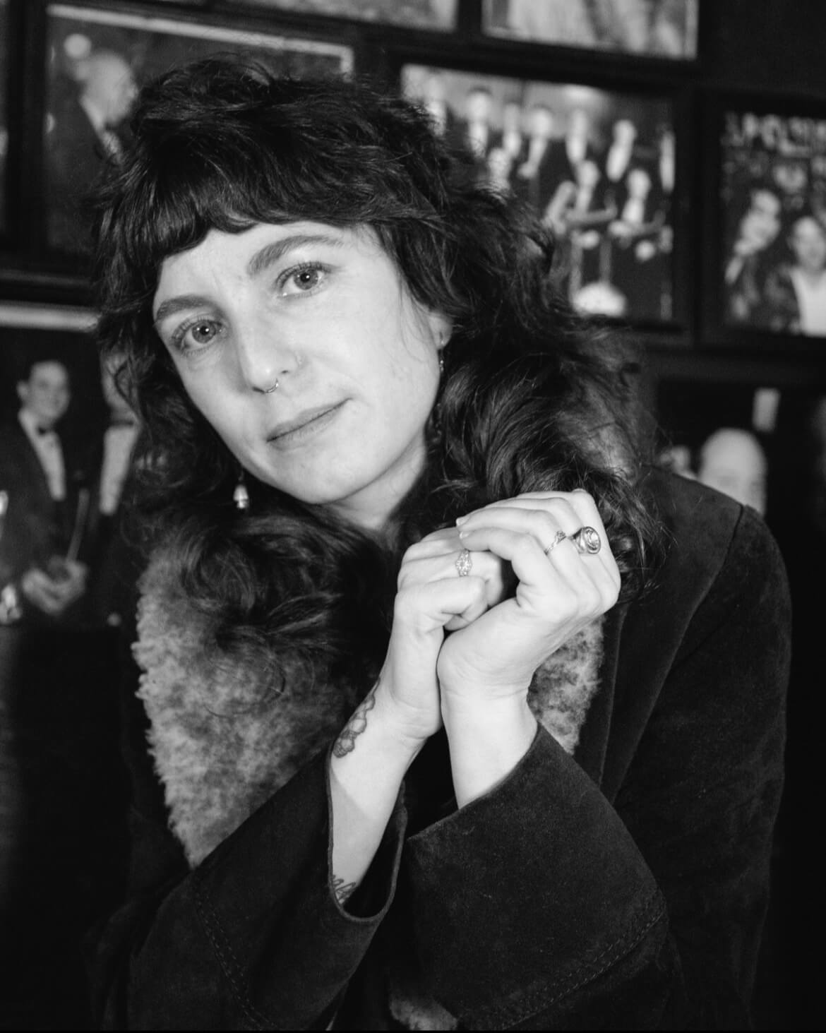 Portrait of Sasha Wortzel in black and white. A white woman with wavy brown hair in a fur trimmed coat and looks at the camera with their hands clasped.