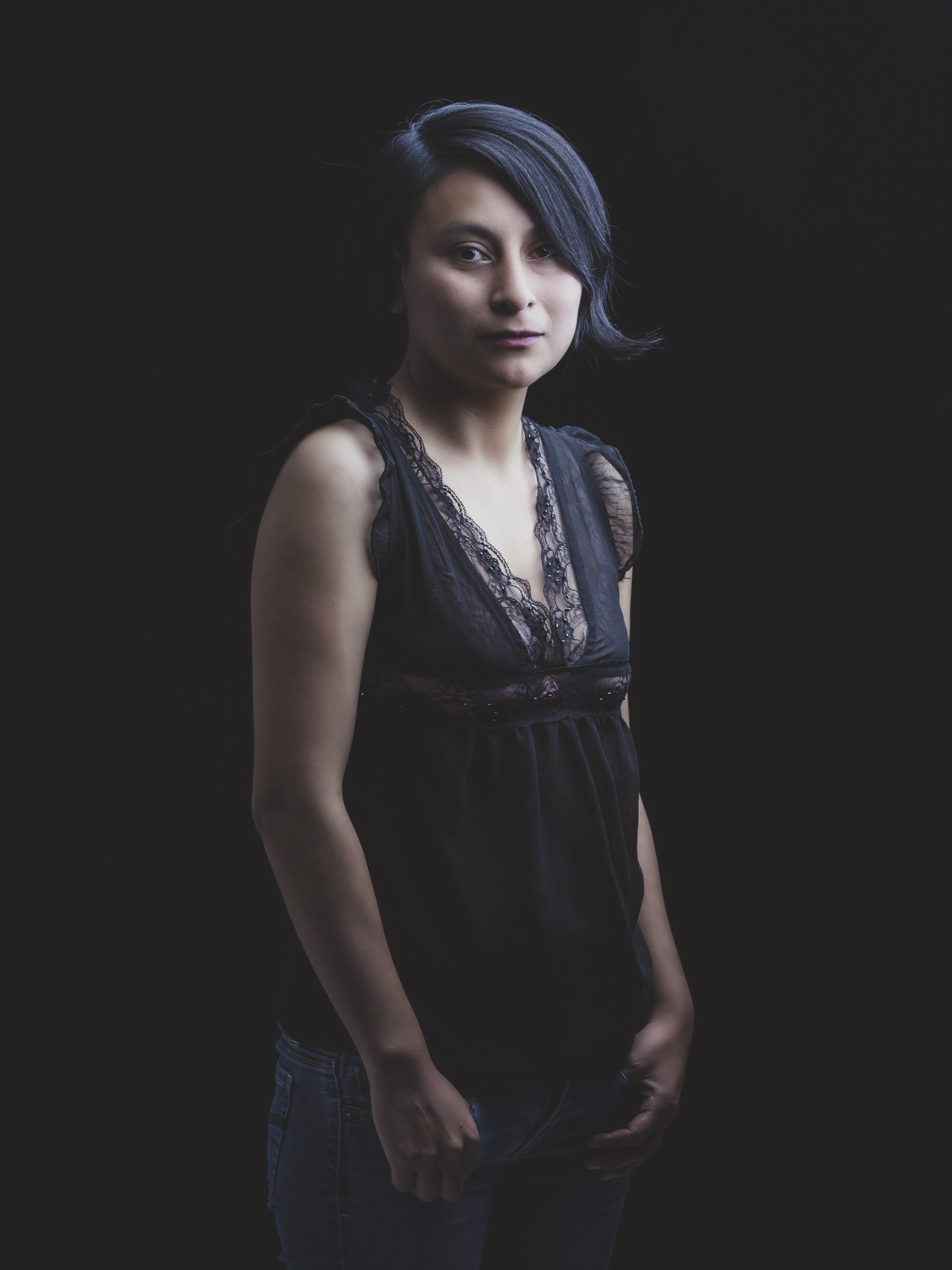 A mid-shot of Rilda Paco, a young Aymara woman standing against a dark background, her expression calm and introspective. She has straight, dark hair that falls just above her shoulders. She wears a sleeveless black top with lace detailing along the neckline, paired with jeans. The lighting is soft, highlighting her features gently, with the contrast between the darkness around her and the lighter tones of her skin and clothing creating a striking, minimalist portrait.