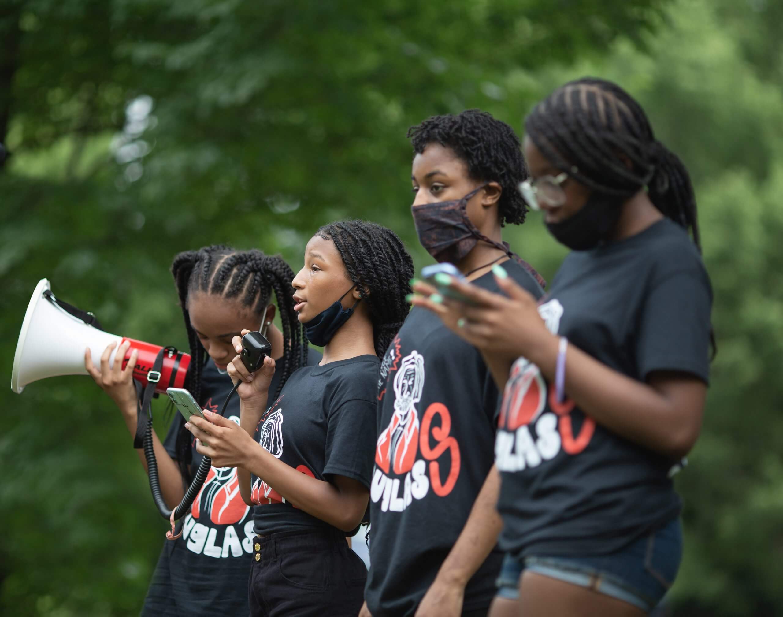 Still from Change The Name. A group of young girls in matching t-shirts and mask speak into a megaphone.