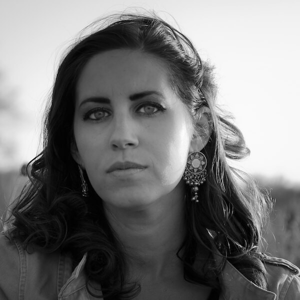 black and white headshot of filmmaker Erika Cohn. a young woman with shoulder length, dark wavy hair looks directly at the camera. She has light colored eyes ringed with dark eyeliner and wears large silver earrings.