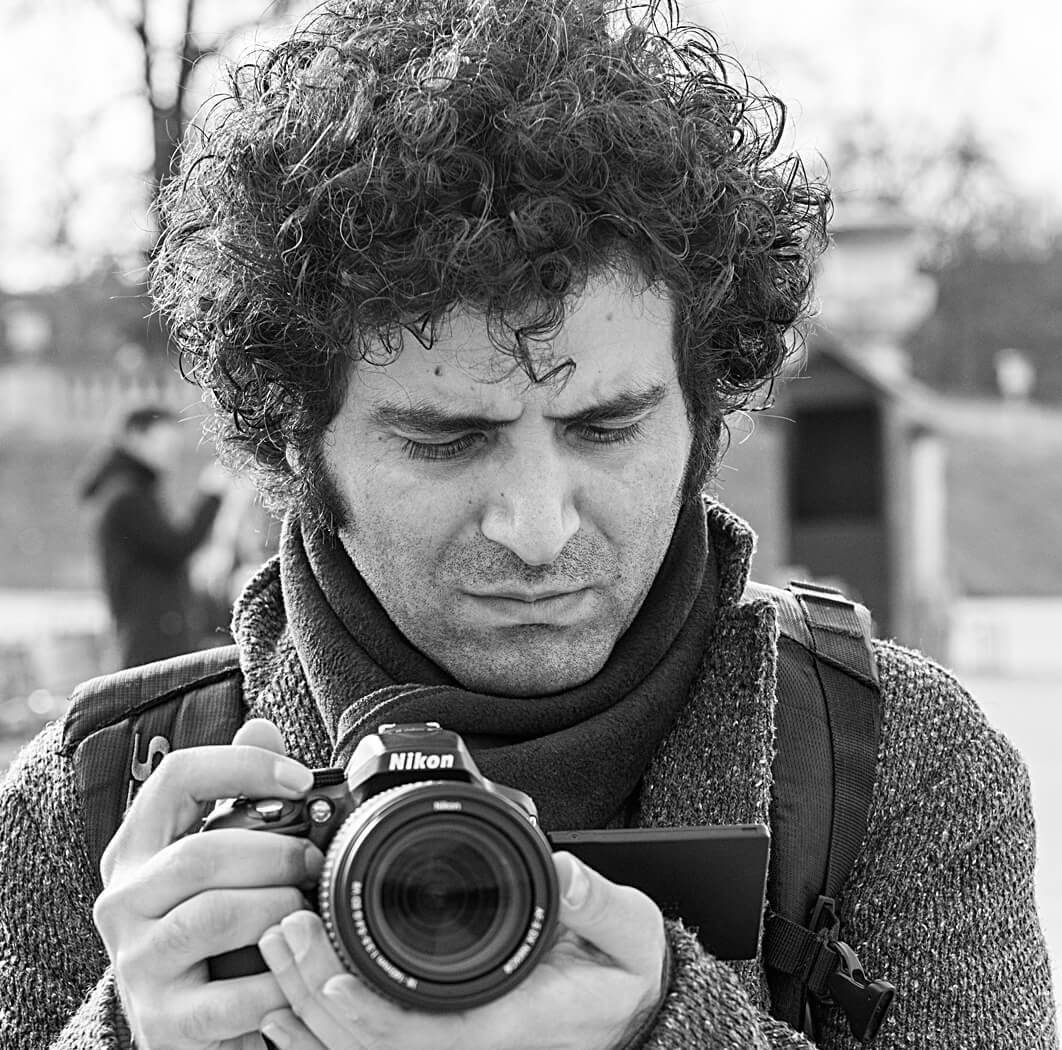 A black and white headshot of Mohammadreza Eyni. He has full curly hair, wears a heavy scarf and coat and holds a digital Nikon camera in his hand. He looks at the screen of the camera, not directly at the viewer. 