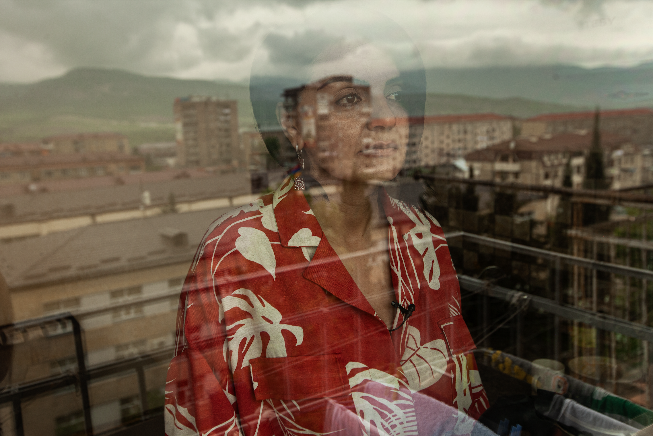 Still from There Was, There Was Not. A woman with dark hair and a red dress stares out a window, the buildings of a city are reflected against her face in the glass.