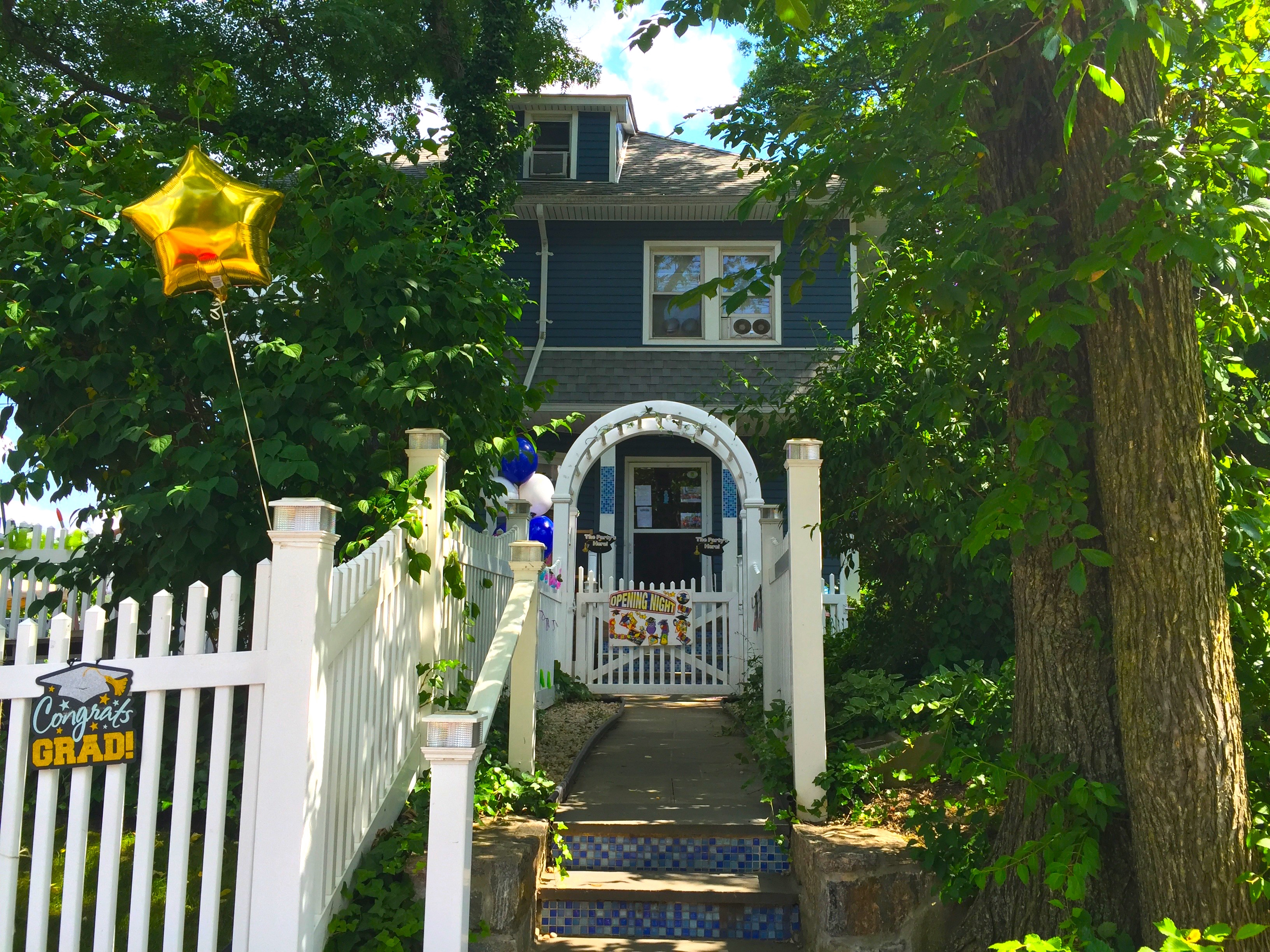 Still from Through the Night. An Exterior shot of a blue house surrounded by green vegetation. There is a balloon in the shape of a golden star tied to the white fence in front of the house.