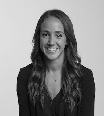 A black and white headshot of Alex Simon. A woman with long, wavy brown hair smiles looking directly at the camera. 