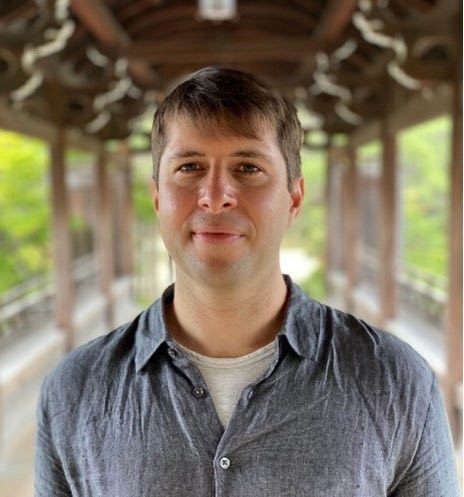 A man with brown hair looks into the camera, smiling, blurry background. 