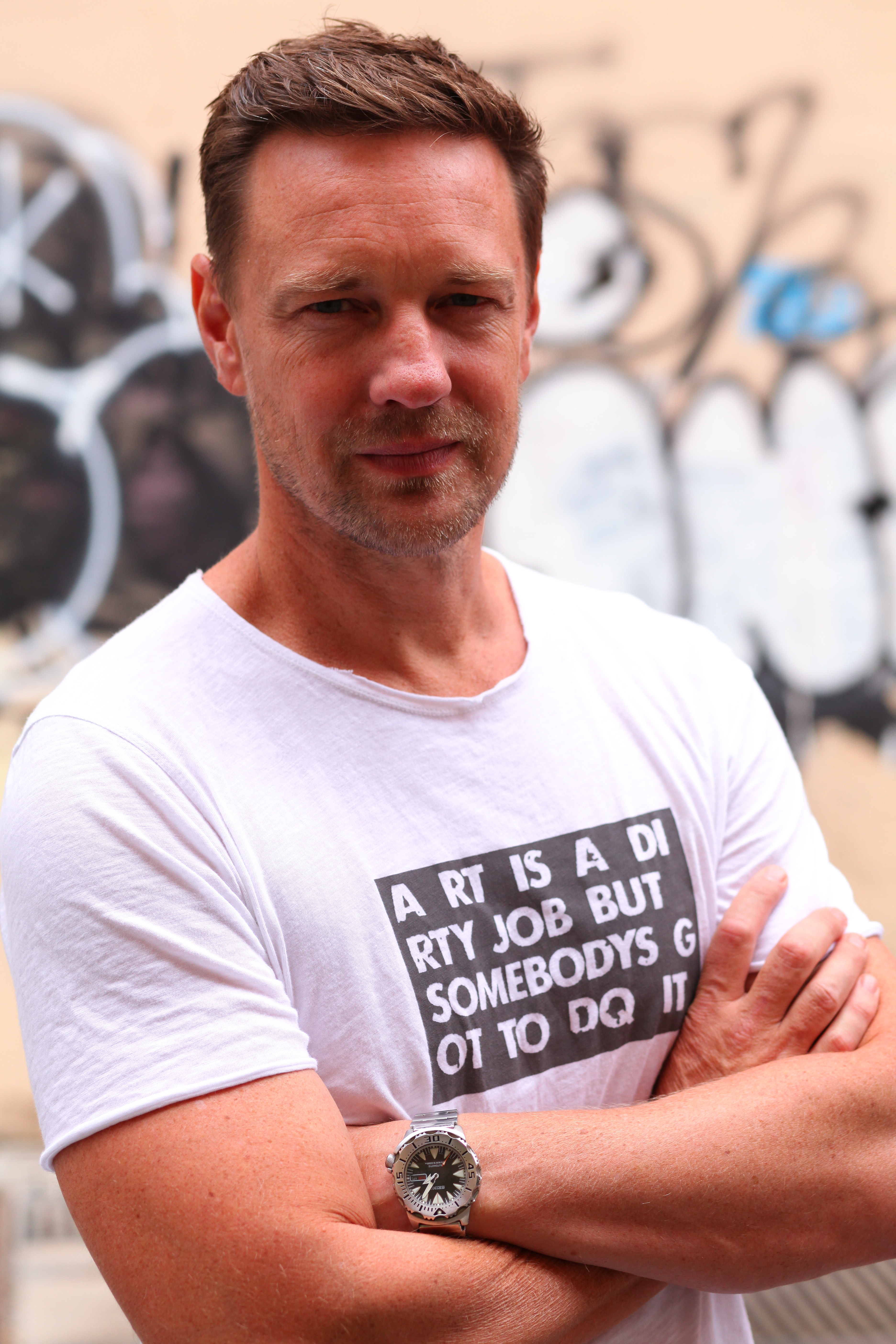 A close up of Dan Fallshaw, a white male with short brown hair and a light beard, standing with his arms crossed. He is wearing a white T-shirt with the text "ART IS A DIRTY JOB BUT SOMEBODY'S GOT TO DO IT" printed in bold letters on the front. Dan has a serious, confident expression on his face. A silver watch is visible on his wrist. The background is blurred, featuring graffiti art, which adds an urban feel to the portrait.