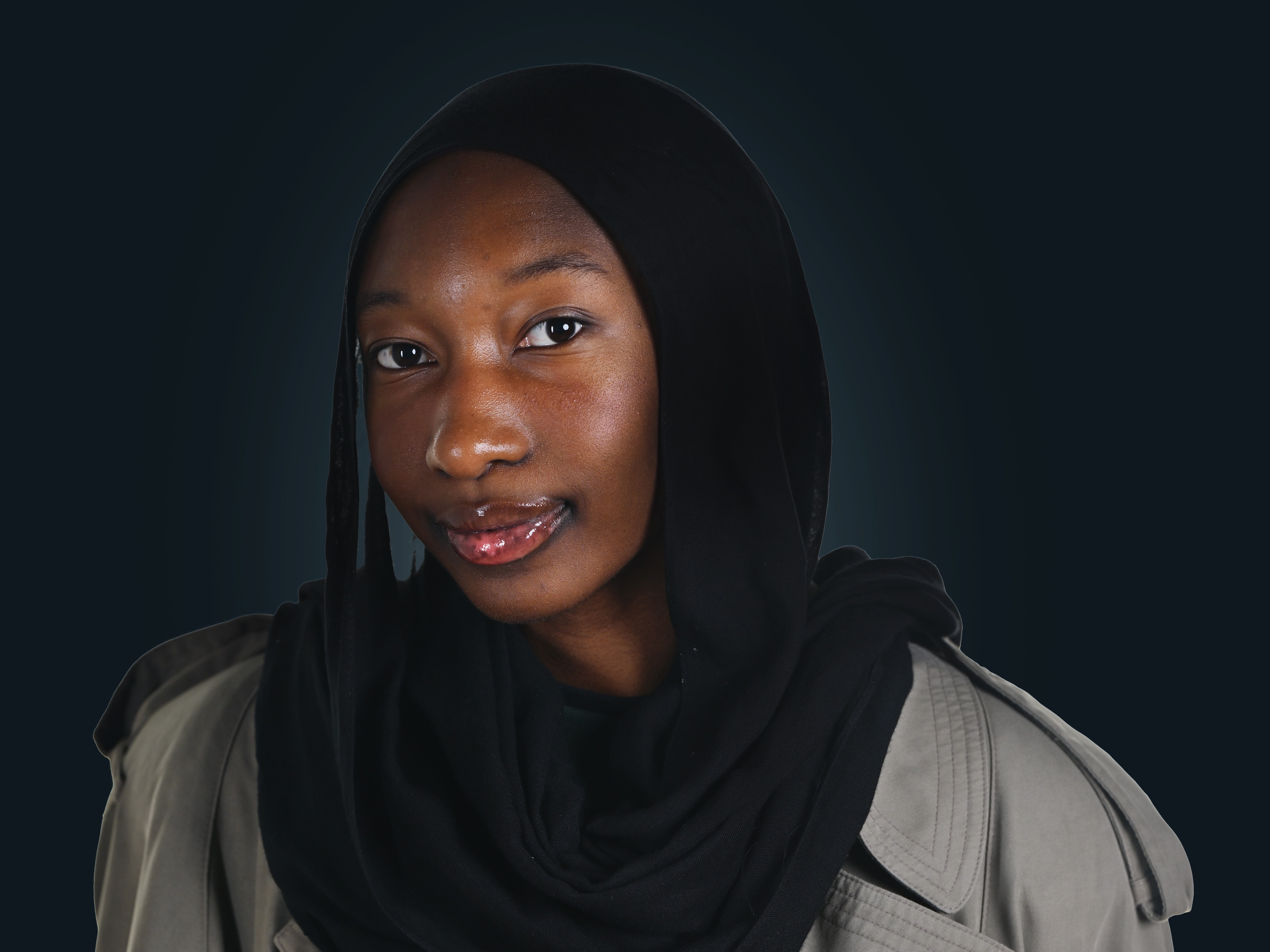 A portrait of a woman wearing a black head wrap and trench coat looking into the camera.