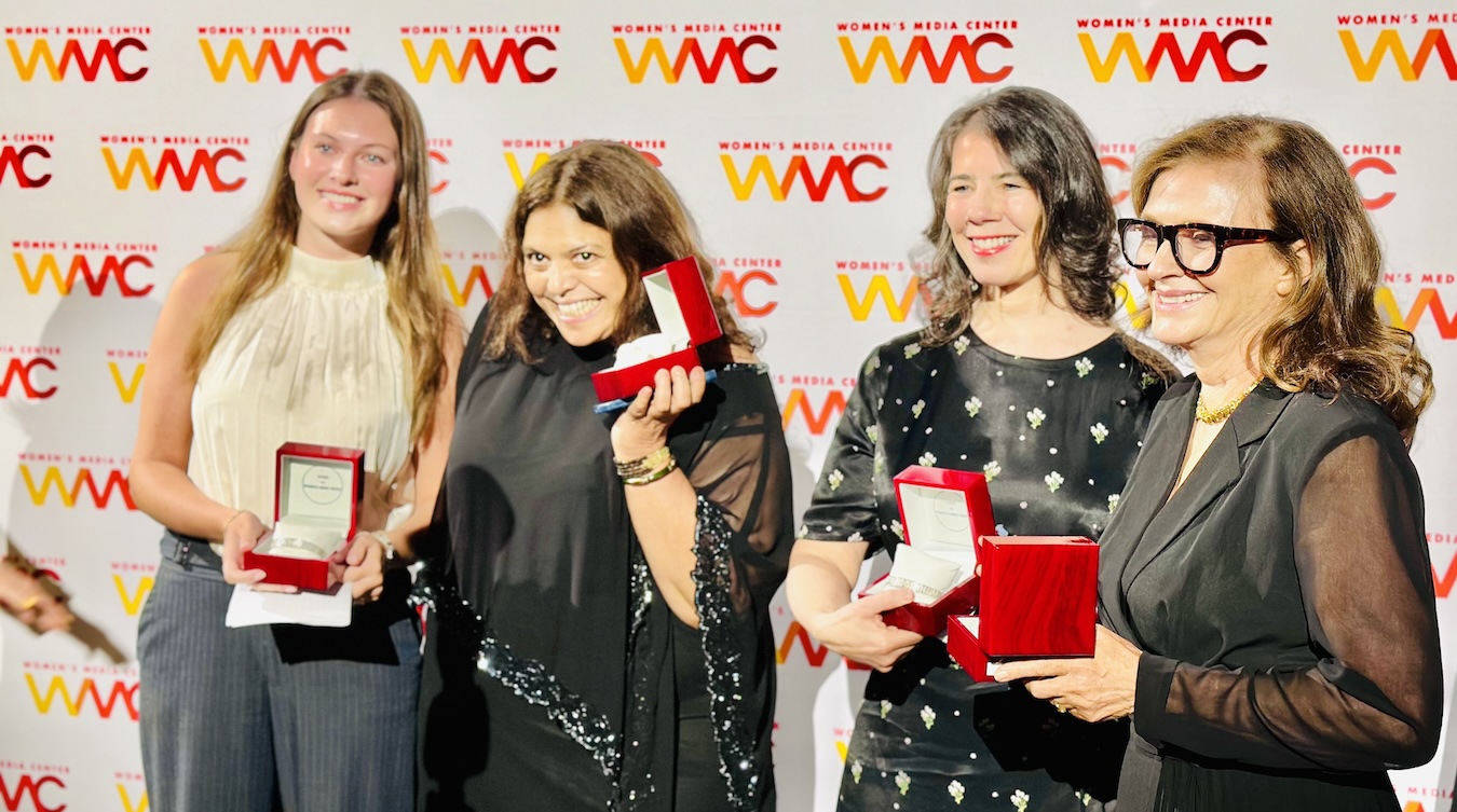 Avery Benello, Judith Helfand, Jenni Wolfson, Wendy Ettinger stand together on the red carpet in front of a step-and-repeat backdrop with the Women's Media Center logo on it. They hold open small red velvet boxes showing their awards for the Pat Mitchell Lifetime Achievement Award