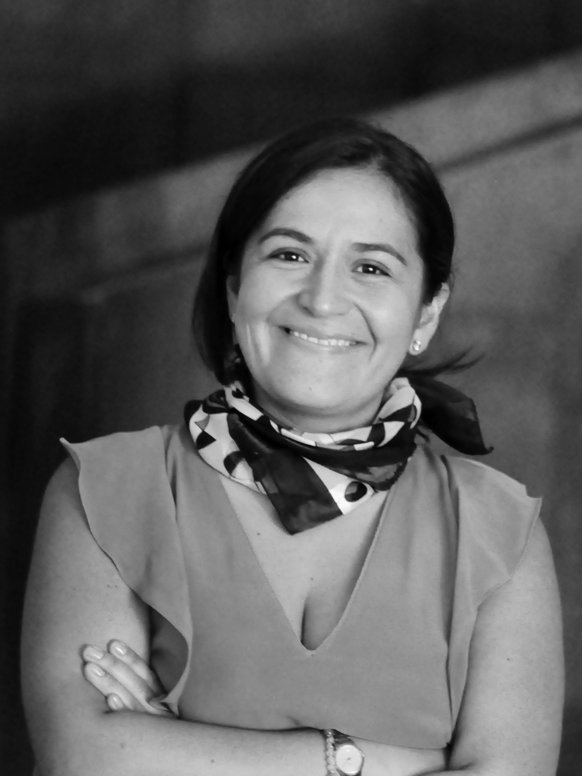 Headshot of a woman smiling, leaning her head to the left. Her hair is tied in the back, she wears a handkerchief on her neck and a sleeveless blouse.